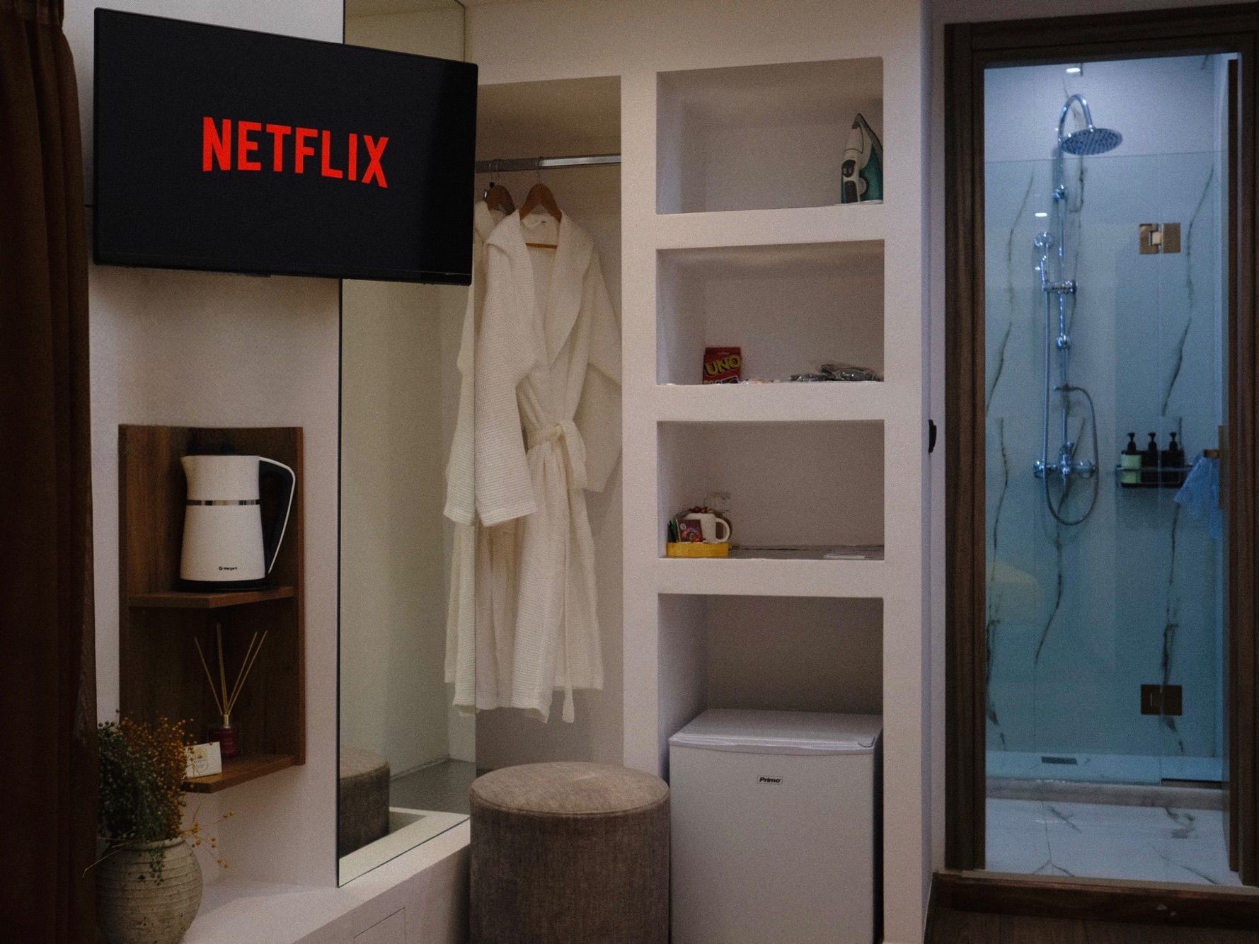 A bathroom with a Netflix sign, a robe hanging, shelves with toiletries, a shower, and a small trash bin.