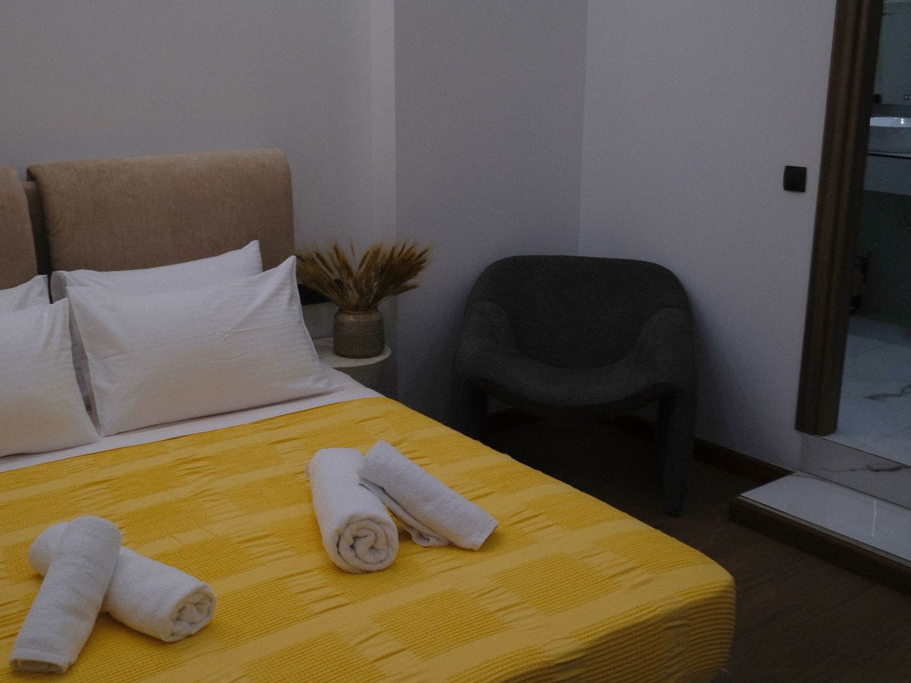 Quarto de hotel com cama, almofadas, uma cadeira e uma pequena mesa com uma planta.