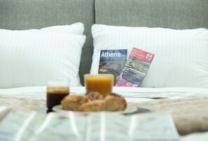Un lit avec des oreillers blancs, un plateau de petit-déjeuner avec des croissants et un verre de jus, et deux guides de voyage posés sur les oreillers.