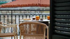 Un balcon avec des fauteuils en osier, une table avec des éléments pour le petit-déjeuner et une vue sur un immeuble aux toits recouverts de tuiles rouges.