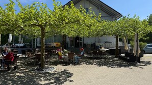 Ein Sitzbereich im Freien mit Tischen und Stühlen, ein Gebäude mit großen Fenstern und eine Pergola mit üppigem Grün.