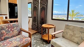 Ein modernes Wohnzimmer mit einer Couch, Sesseln, einem kleinen runden Tisch und einem Fernseher. Große Fenster bieten Blick auf das Meer und Boote.