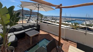 Eine Terrasse mit Sofa, Tisch, Sonnenschirm und Blick auf einen Yachthafen.