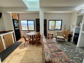 A room with a wooden dining table, a sofa, and two windows with a view of the beach.