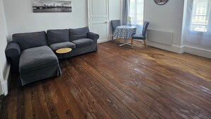 Ein Wohnzimmer mit grauem Sofa, einem kleinen runden Tisch, einem Sessel, einer Uhr und einem Fenster mit Vorhängen.