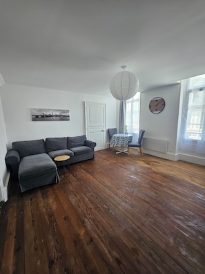 Een woonkamer met een grijze bank, een klein rond tafeltje, een stoel, een klok en een raam met gordijnen.