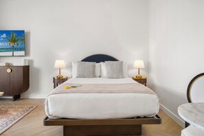 Ein ordentlich eingerichtetes Schlafzimmer mit einem Bett, Nachttischlampen, einem Nachttisch, einem Zierkissen, einem kleinen Tisch und einem an der Wand montierten Fernseher.