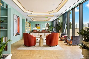 Eine geräumige Lobby mit hoher Decke, einer zentralen Sitzgruppe mit zwei roten Sesseln, einem Couchtisch und einer Couch. Große Fenster bieten Blick auf die Stadtlandschaft.
