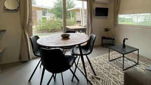 Una habitación con una mesa redonda, sillas negras, un televisor pequeño, una planta en maceta y una ventana con cortinas.