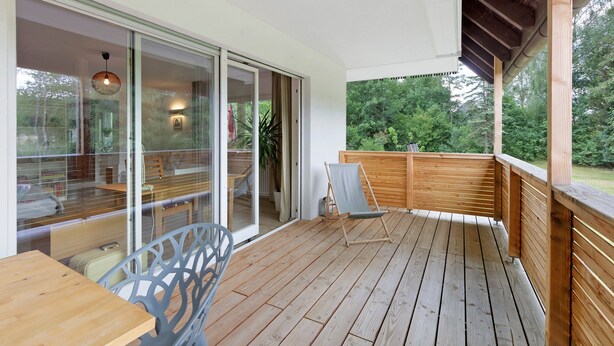 Una terraza de madera con una mesa, sillas, un banco de madera y un puerta de cristal que da a un espacio interior con una silla y una mesa.
