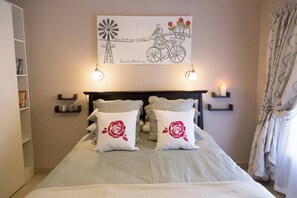 A neatly made bed with two rose-patterned pillows, a wall-mounted artwork depicting a windmill and a bicycle, and bedside tables with candles. - Serene Holiday Retreat Near Hartbeespoort Dam (Hartbeespoort)
