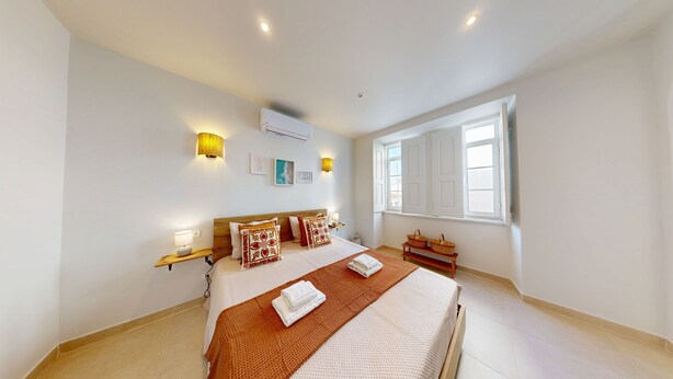 A hotel room with a large bed, two bedside tables, a window with shutters, and wall-mounted lighting.