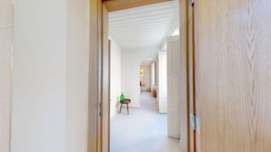 A hallway with a door at the end leading to a room with a bed and a small table.