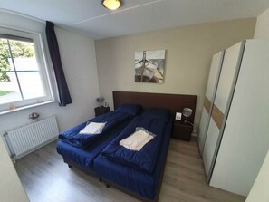 Ein Schlafzimmer mit zwei Betten, einem Fenster, einem Heizkörper, einem Nachttisch, einer Lampe und einem Kleiderschrank.