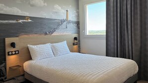 Una habitación de hotel con cama, cabecero con interruptor de luz, toma de corriente en la pared y ventana con cortinas.