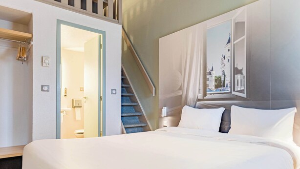 A hotel room with a large bed, a view of a castle through a window, a staircase, and a door leading to a bathroom.