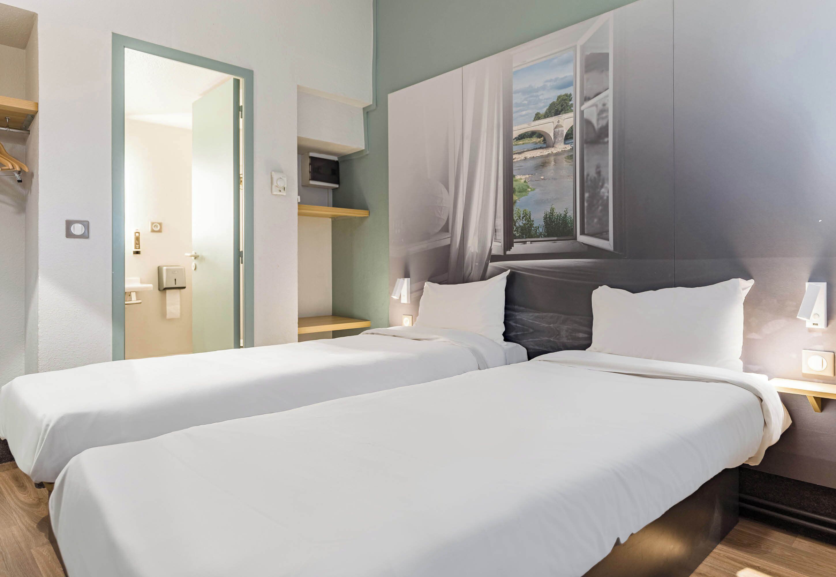 Habitación de hotel con dos camas, una ventana con vista a un puente, un espejo y una puerta que da a otra habitación.