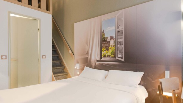 Une chambre à coucher avec un lit, une fenêtre avec des rideaux, un escalier et un luminaire fixé au mur.