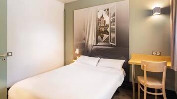 A hotel room with a bed, a desk, and a chair. There is a painting on the wall above the bed.