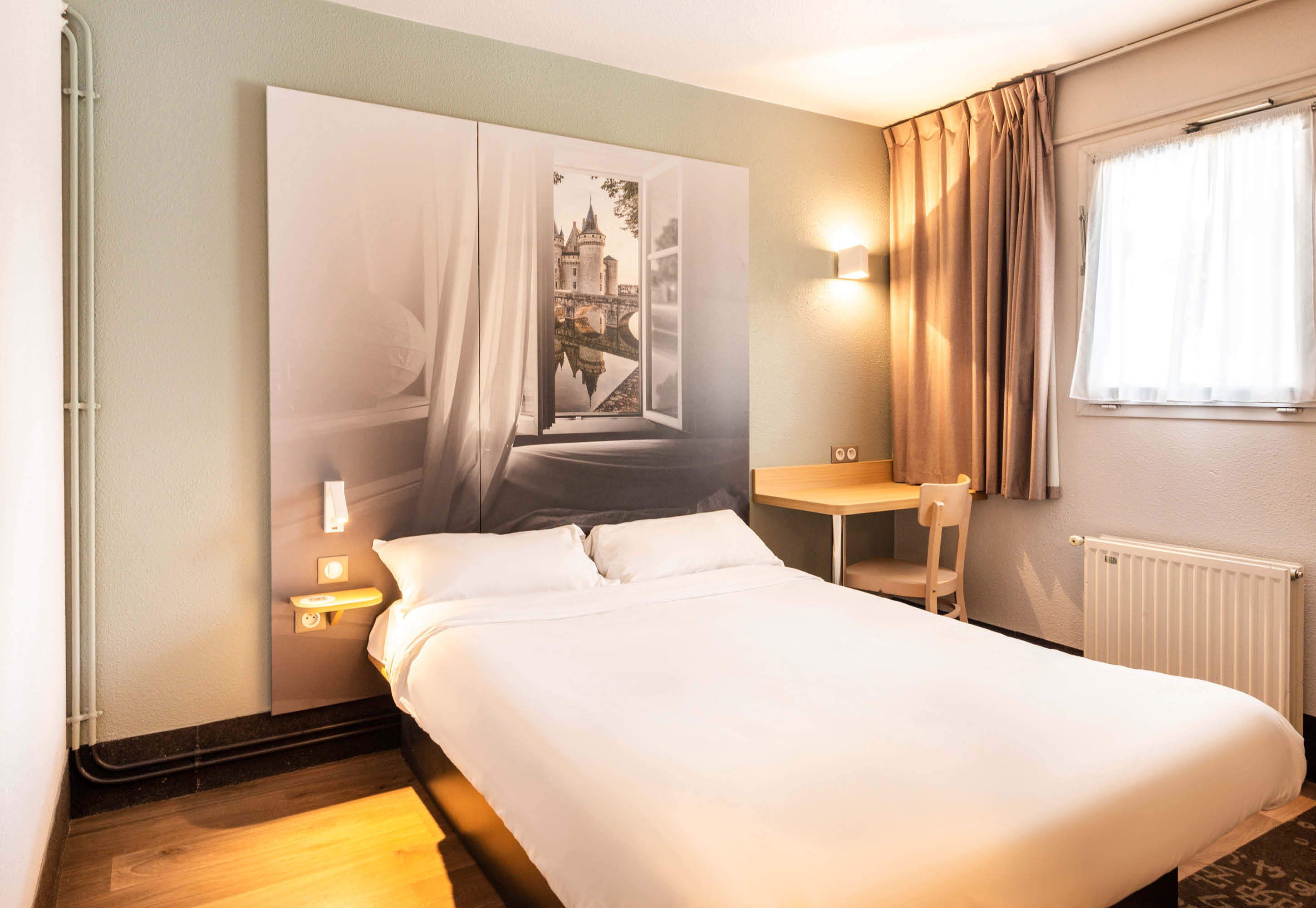 Een hotelkamer met een bed, nachtkastje, stoel, raam met gordijnen en een wandlamp.
