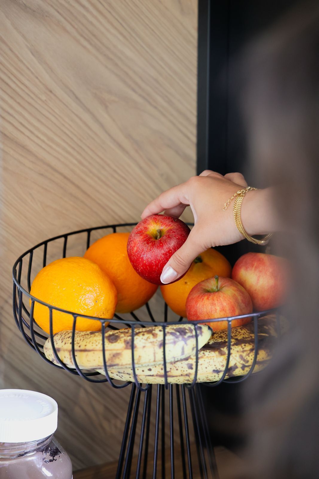 Una mano alcanza una manzana en una cesta de mimbre con naranjas y plátanos.