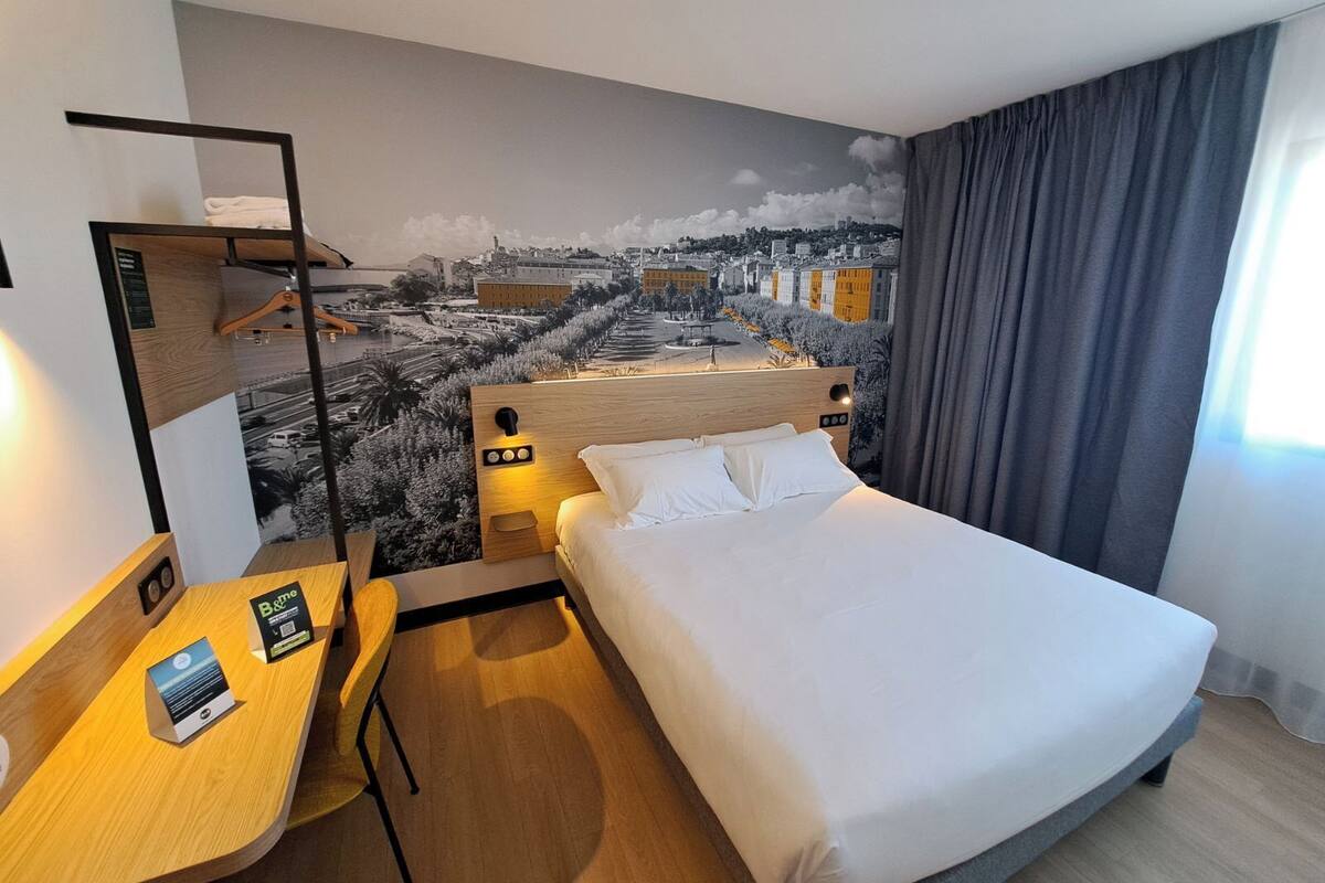 A hotel room with a bed, a desk, a chair, a nightstand, a mirror, and a cityscape mural on the wall.