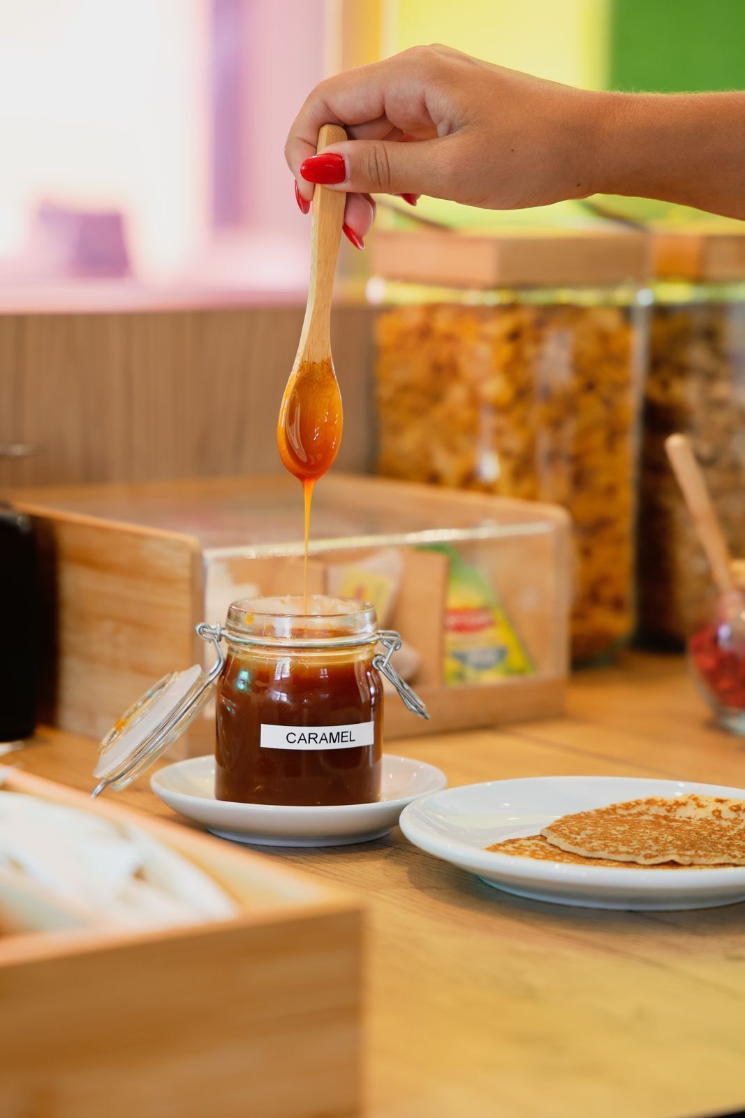 Una mano sosteniendo una cuchara sobre un tarro etiquetado 'CARAMEL' con un plato de panqueques cerca.