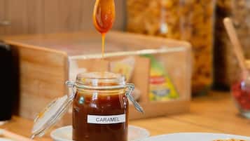 Una mano sosteniendo una cuchara sobre un tarro etiquetado 'CARAMEL' con un plato de panqueques cerca.