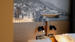 Quarto de hotel com uma cama, roupa de cama branca, cabeceira com luzes de leitura fixadas na parede e um grande mural com vista para a cidade.