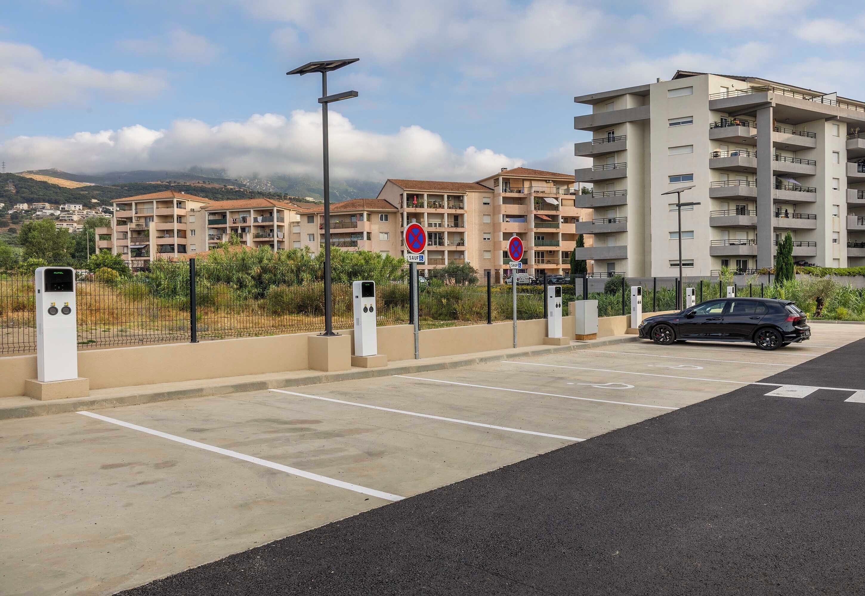Un espace de stationnement équipé de bornes de recharge pour véhicules électriques, un panneau indiquant l’interdiction de se garer, et un immeuble résidentiel à plusieurs étages en arrière-plan.