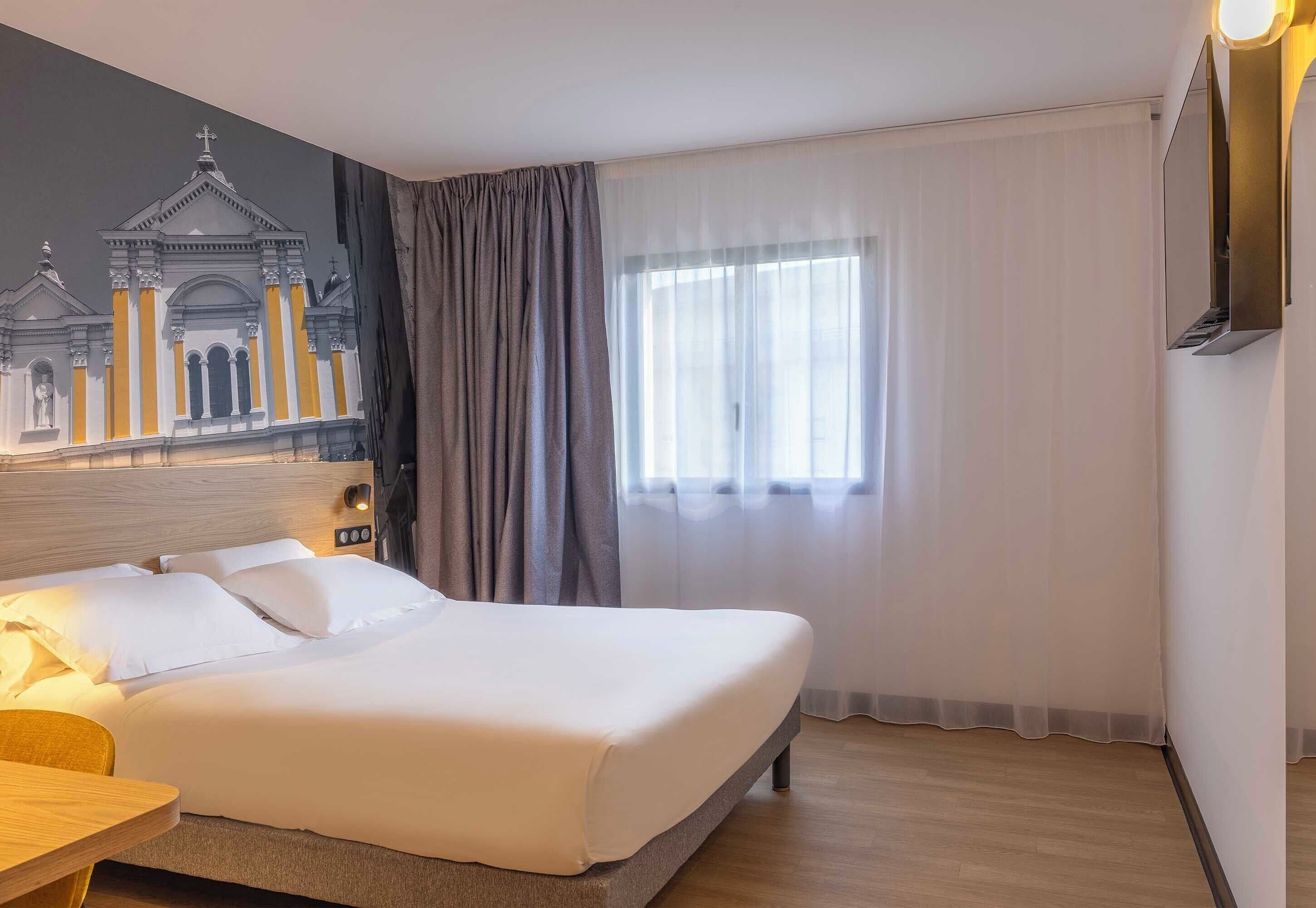 A hotel room with a bed, a nightstand, a wall-mounted lamp, a window with curtains, and a mural of a building.