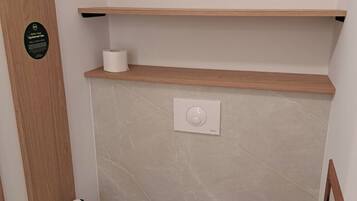 Un baño pequeño con un inodoro, un estante de madera, un dispensador de papel higiénico y un botón de descarga.