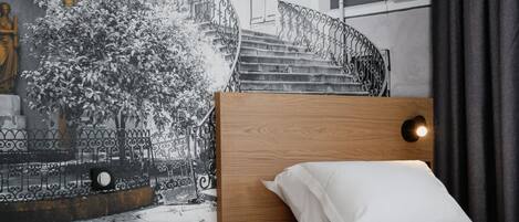A bedroom with a bed, a headboard, a nightstand, a wall-mounted lamp, and a window with curtains.