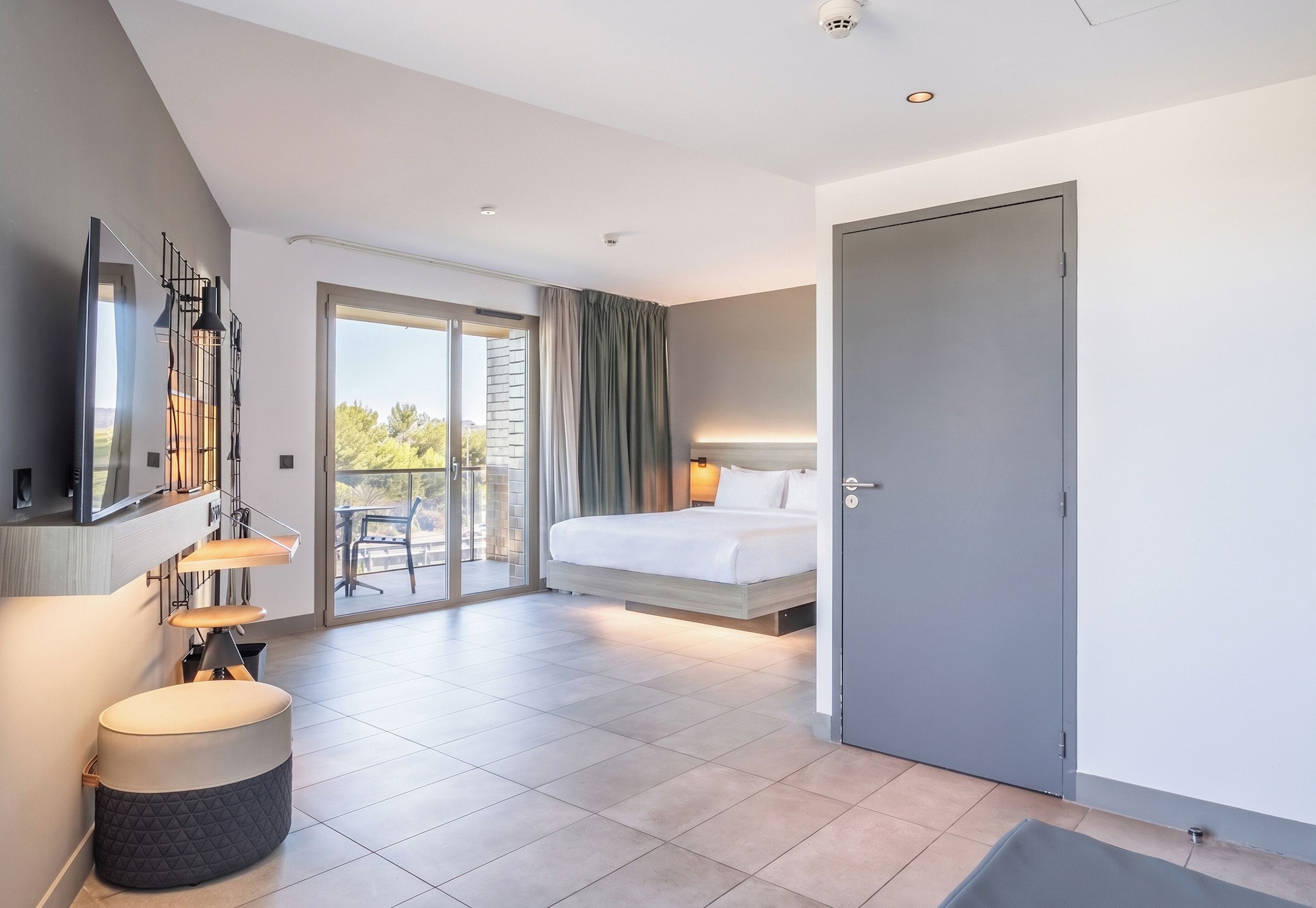 A modern hotel room with a bed, a seating area with a table and chairs, a large window, and a door.