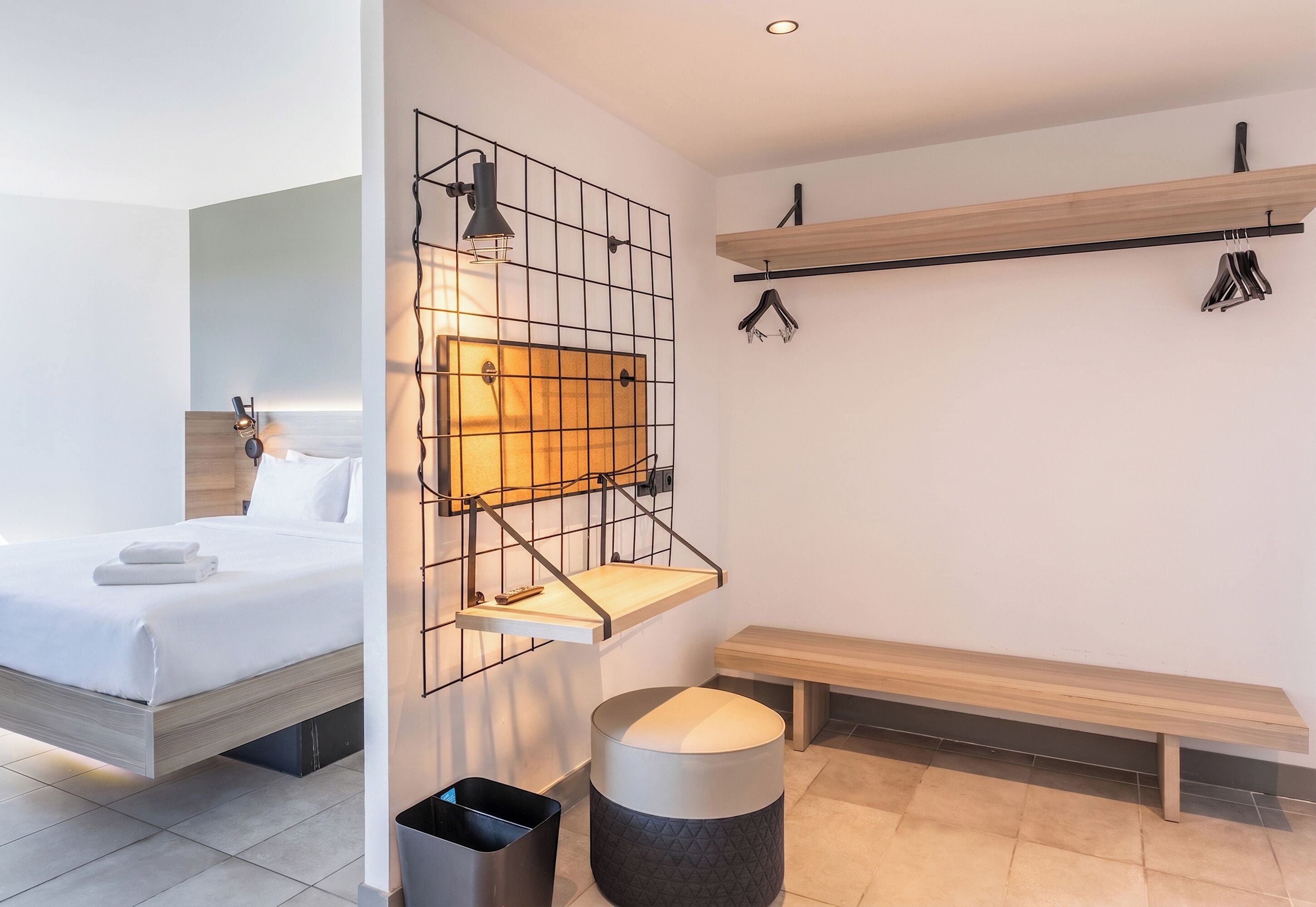 Et moderne hotelværelse med seng, vægmonteret hylde, bænk og skraldespand.