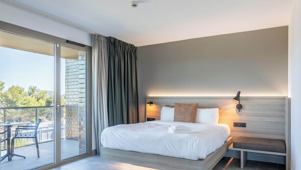A modern hotel room with a large bed, a bedside table, a chair, a sliding glass door to a balcony, and a wall-mounted light fixture.