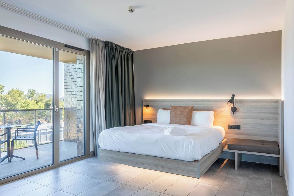 Una habitación de hotel moderna con una cama grande, una mesita de noche, una silla, un ventanal corredero a un balcón y una lámpara fijada a la pared.