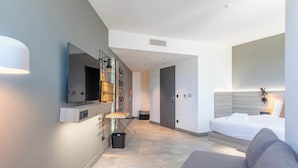 A modern hotel room with a bed, a sofa, a desk with a chair, a mirror, and a television.