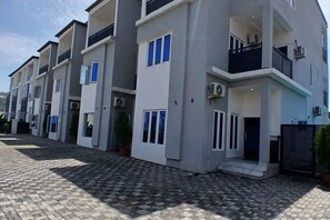 Un immeuble résidentiel moderne à plusieurs étages, doté de balcons, de climatiseurs et d’une allée pavée.