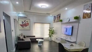 Una sala de estar moderna con un sofá, una mesa de comedor con sillas, un televisor en la pared y un ventanal con persianas.