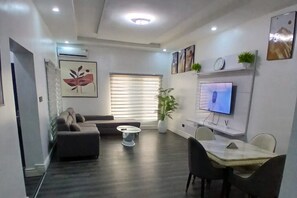 Un salon moderne avec un canapé, une table à manger et des chaises, un téléviseur fixé au mur et une grande fenêtre avec des stores.