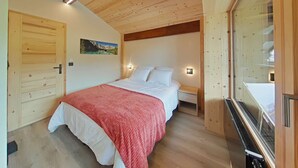 Un dormitorio con techo de madera, una cama con ropa de cama blanca, una manta roja, una mesita de noche, una lámpara de pared, un armario de madera y una ventana con vistas.