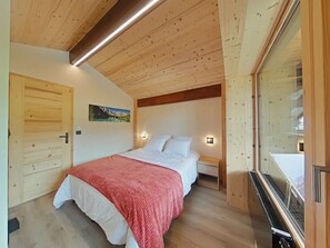 Ein Schlafzimmer mit Holzbalkendecke, einem Bett mit weißer Bettwäsche, einer roten Decke, einem Nachttisch, einer Wandleuchte, einem hölzernen Kleiderschrank und einem Fenster mit Aussicht.
