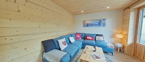 A cozy cabin interior with a wooden ceiling, a blue sectional sofa, a small table, and a painting on the wall.