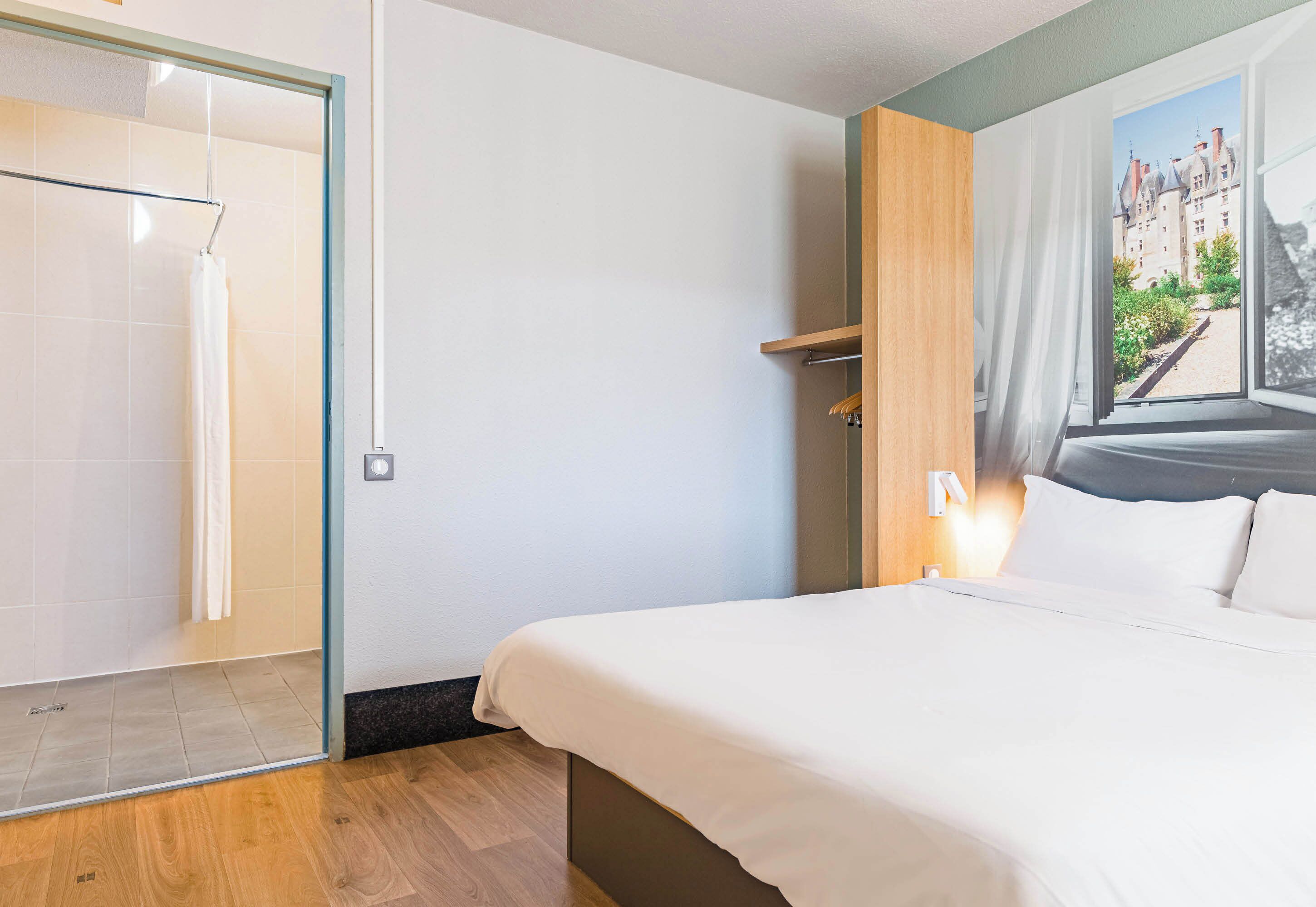 Une chambre d’hôtel avec un lit, une douche, un plancher en bois et une vue sur un immeuble par la fenêtre.