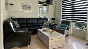Una sala de estar con un sofá de cuero negro, una mesa de centro de madera, una mesita auxiliar y una silla. Hay un cuadro en la pared y una ventana con persianas.