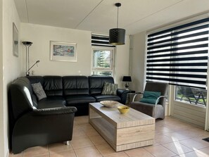 Ein Wohnzimmer mit einem schwarzen Ledersofa, einem Holztisch, einem Beistelltisch und einem Sessel. An der Wand hängt ein Gemälde und ein Fenster mit Jalousien.