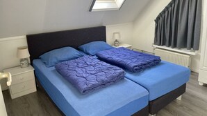 Un dormitorio con tragaluz, dos camas con ropa de cama azul, una mesita de noche con lámpara y una ventana con cortinas.
