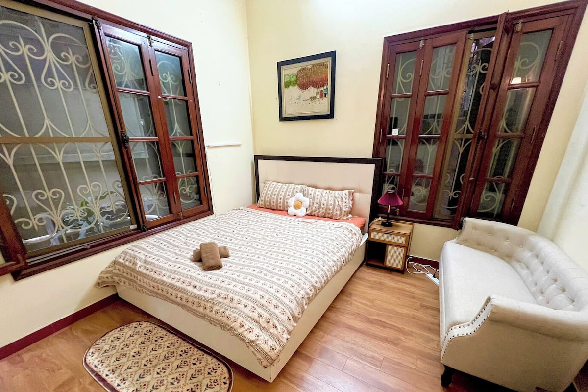 Un dormitorio con cama, lámpara de noche, una silla, alfombra y rejas decorativas en la ventana.