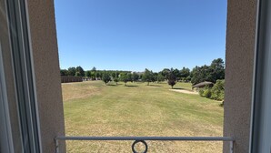 Una vista attraverso una finestra con ringhiera in metallo, un'area verdeggiante, alberi e un edificio sullo sfondo.